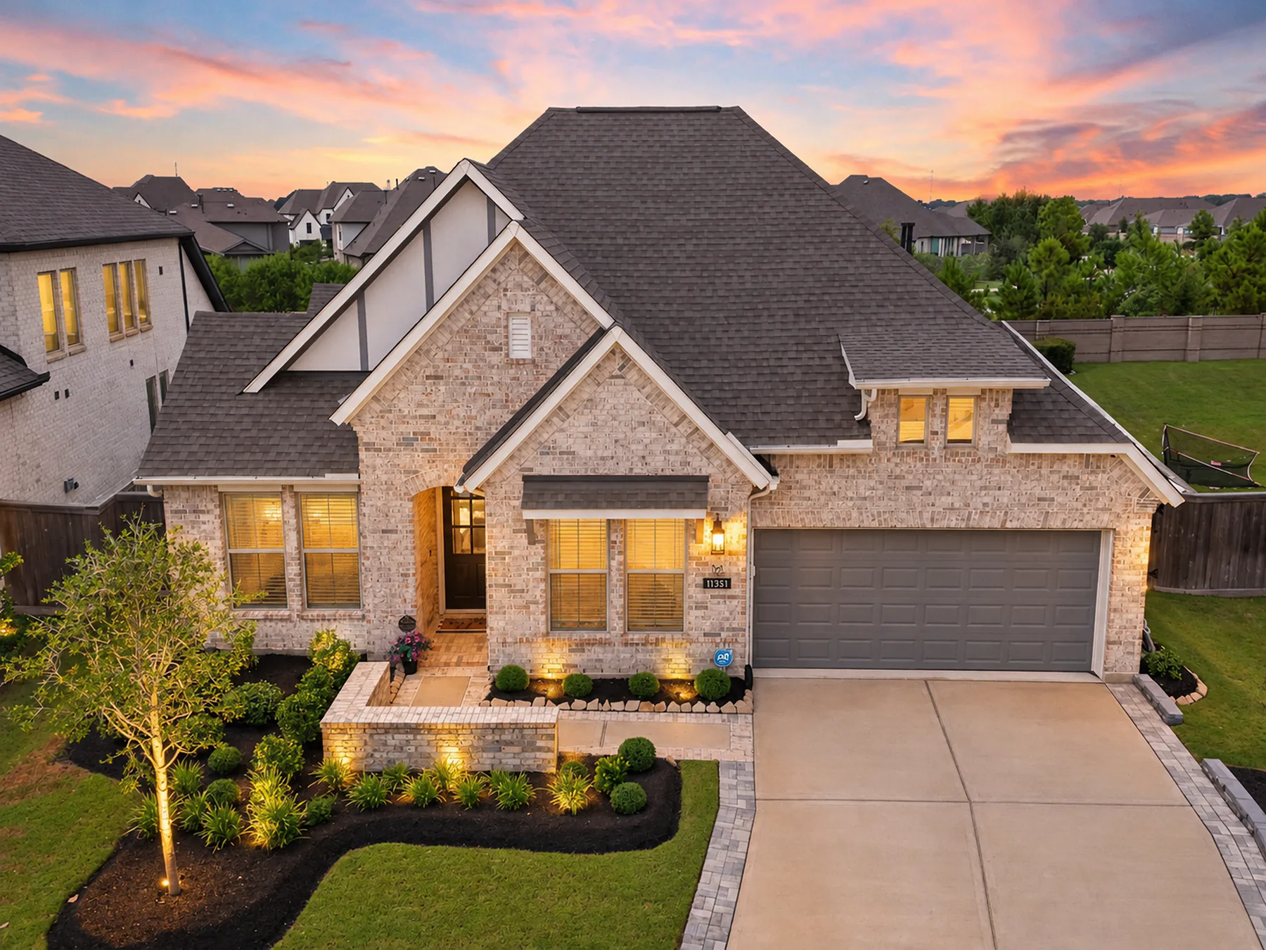 Aerial view of a brick home at sunset with warm windows and landscape lighting